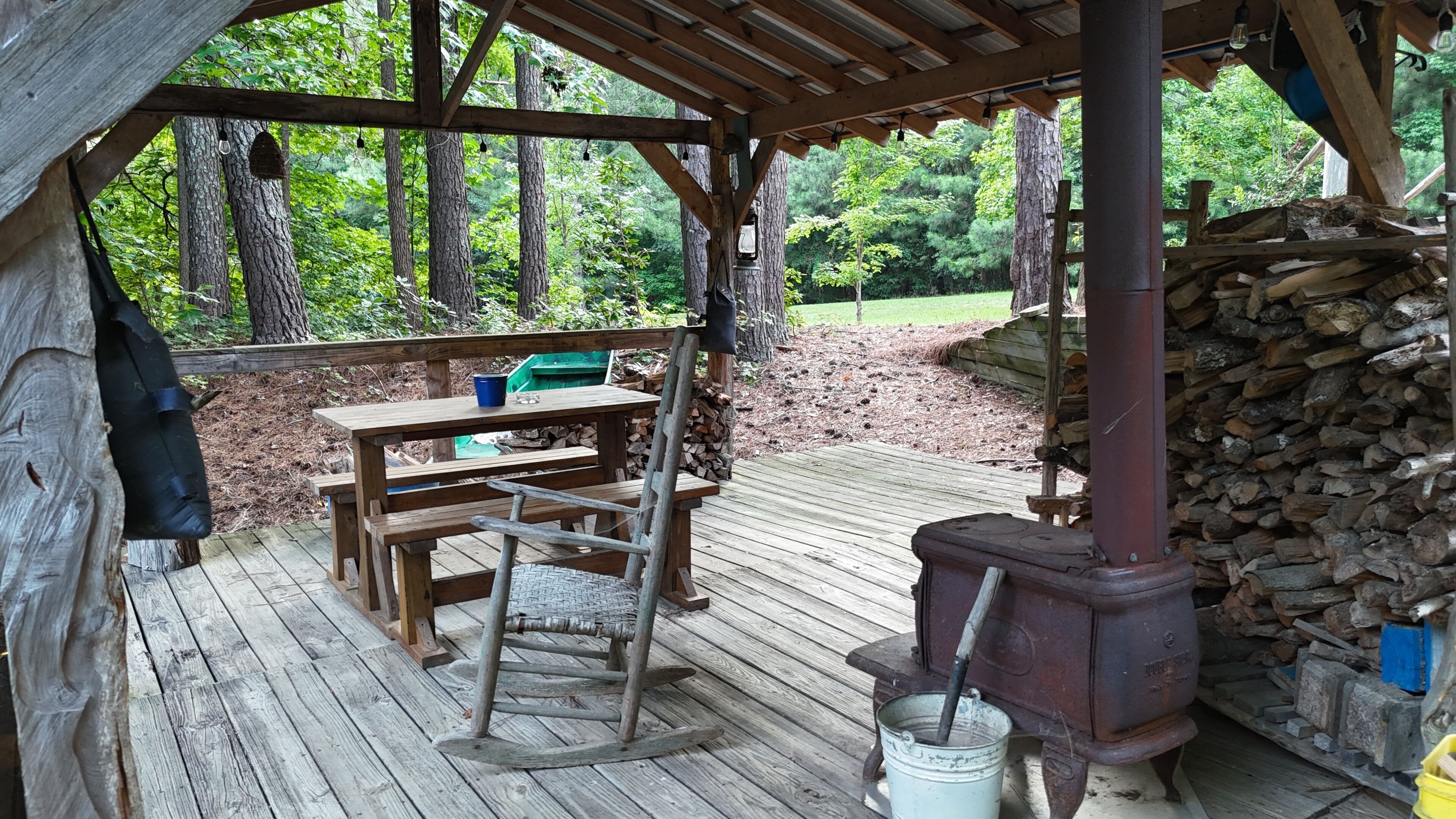 Una terraza de madera al aire libre con una mesa de picnic rústica y bancos, rodeada de árboles.