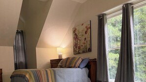 Un dormitorio con cama, mesita de noche, lámpara, ventana con cortinas y un cuadro en la pared.