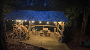Una terraza de madera con un área de picnic cubierta, bancos y una pila de leña.