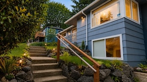Une maison moderne avec un escalier en pierre menant à une terrasse en bois, le tout entouré de verdure.