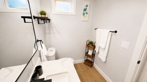 Un baño con lavamanos, inodoro y zona de ducha blancos. Hay dos estantes negros con plantas y una toalla blanca colgada en un perchero.