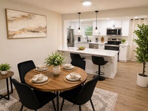 Une cuisine moderne avec des meubles blancs, des appareils en acier inoxydable et des chaises noires disposées autour d’une table à manger en bois.