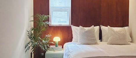 A hotel room with a bed, bedside lamp, a small table with a plant, and a window with a view.