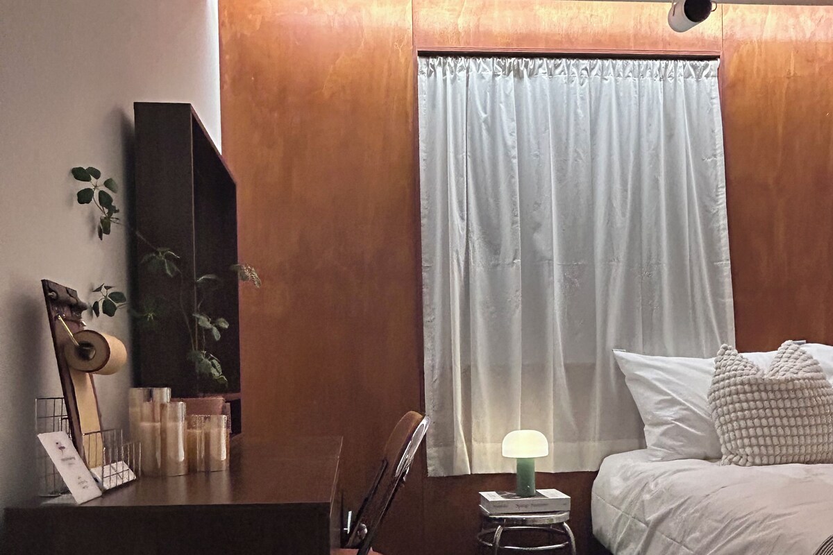 A hotel room with a bed, a desk, a chair, a lamp, and a window with curtains.