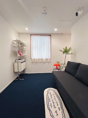 A small, modern living room with a dark blue carpet, a grey sofa, a white shelf unit, and a window with white curtains. - STAY STAMP (Seoul)