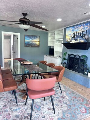 A dining area with a glass table, brown chairs, a ceiling fan, and a coastal mural.