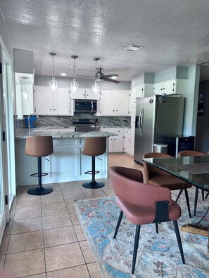 A modern kitchen with a central island, bar stools, a dining table, and a ceiling fan.
