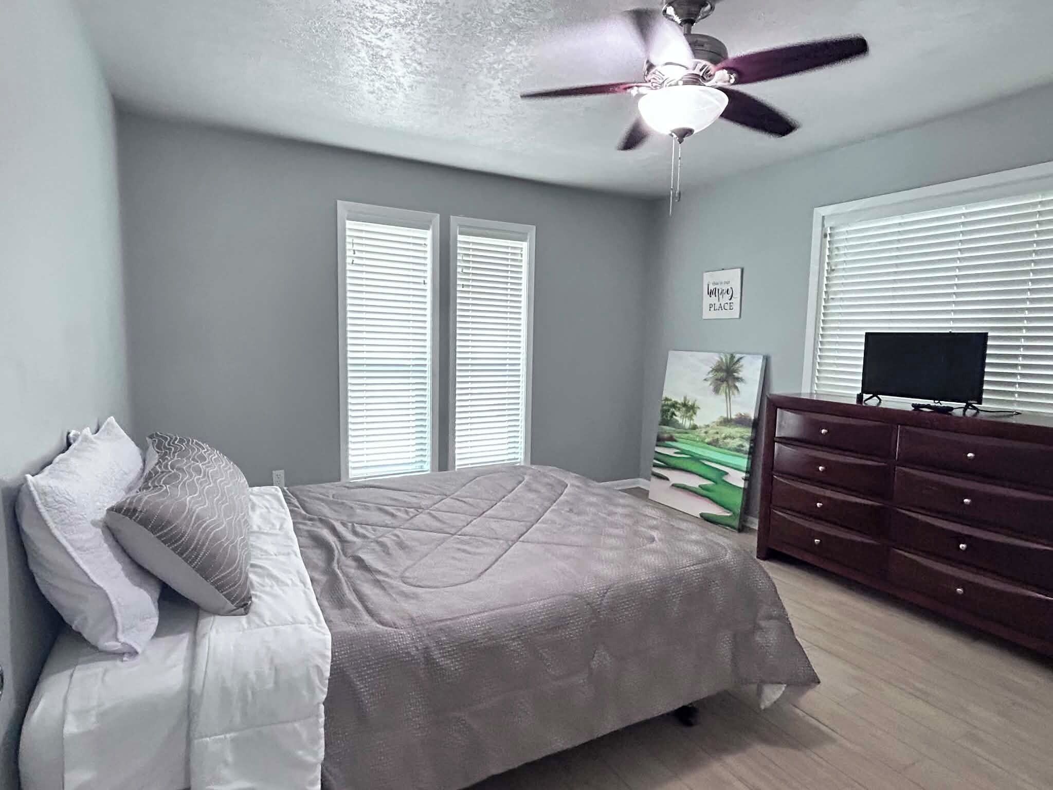 Une chambre à coucher avec un lit, une commode, un ventilateur de plafond et un tableau accroché au mur.