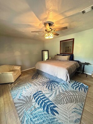 A bedroom with a bed, a chair, a ceiling fan, and a patterned rug.