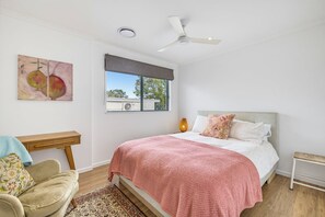 Een slaapkamer met een bed, een plafondventilator, een raam met uitzicht, een schilderij en een nachtkastje.