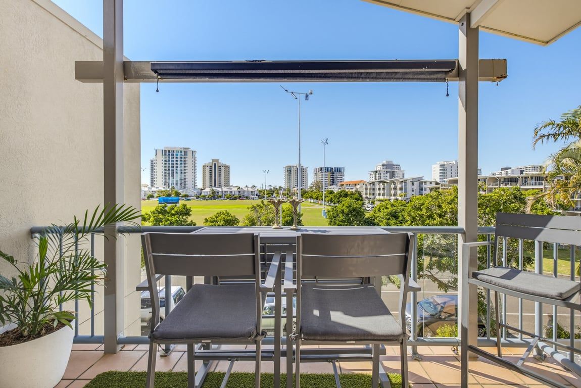 A balcony with two chairs, a potted plant, and a view of a cityscape with buildings and a park.