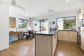 A modern kitchen with wooden flooring, a central island, and large windows offering views of palm trees. - 14 Seashapes - Coastal Comfort With Open Spaces & Beach Glimpses (Maroochydore)
