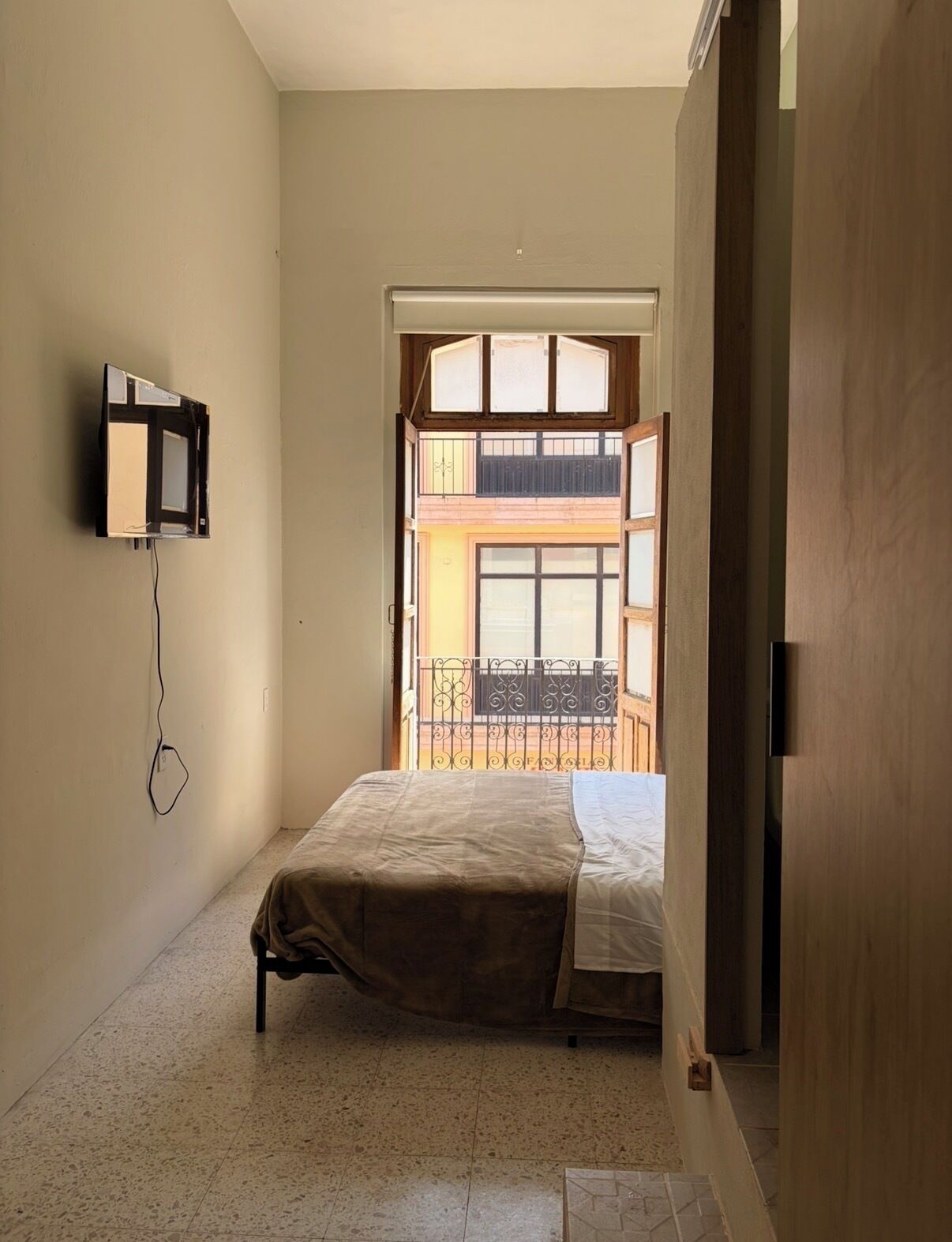 A small, narrow room with a bed, a wall-mounted TV, and a view of an outdoor balcony.