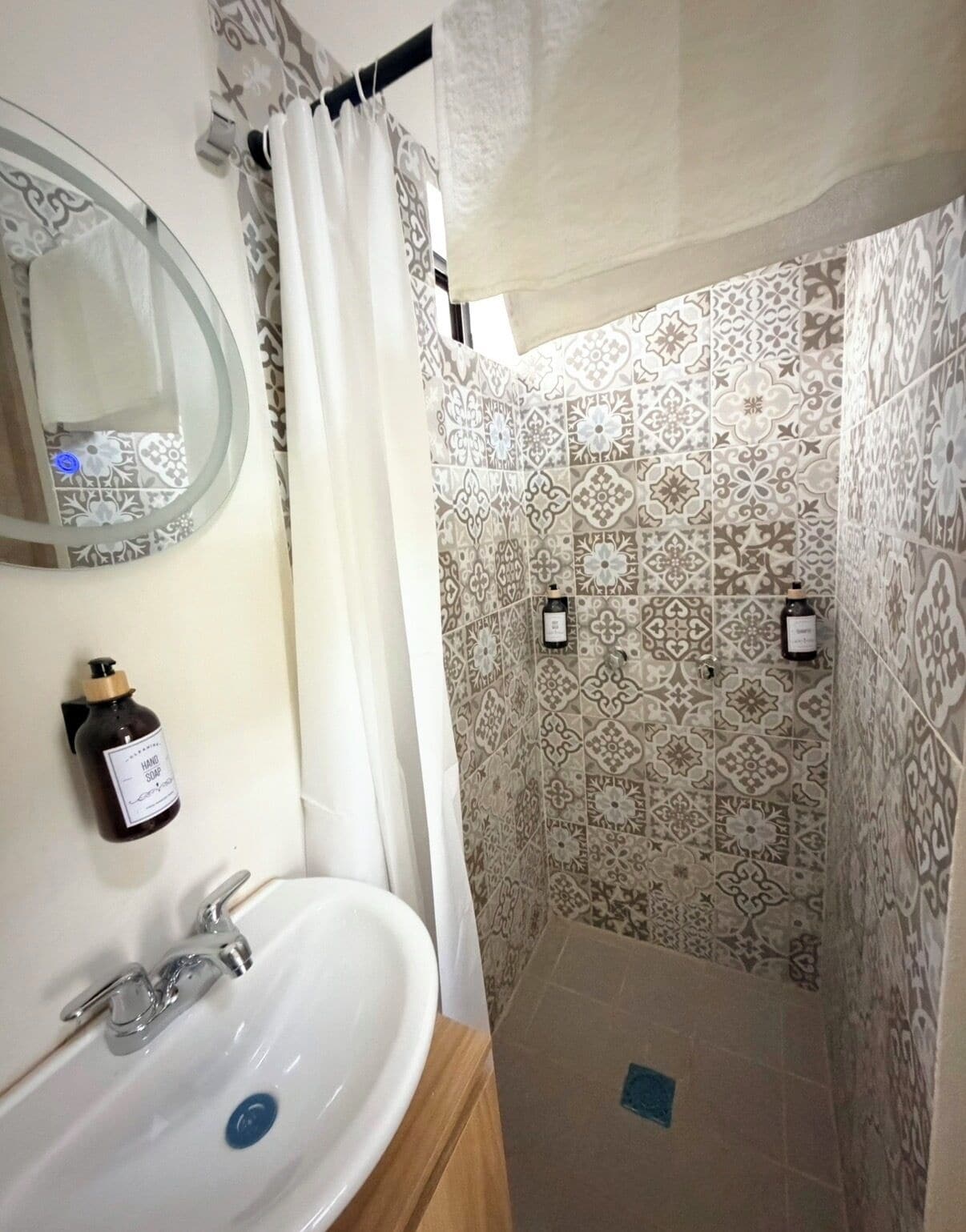 Une salle de bain avec un lavabo blanc, un miroir, une douche aux carreaux décorés et un flacon de savon pour les mains.