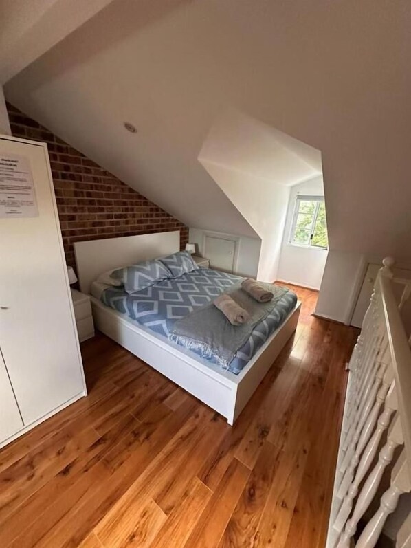 A bedroom with a sloped ceiling, a bed with patterned bedding, a white wardrobe, and a window providing natural light.