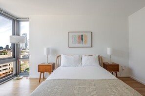 Ein Schlafzimmer mit großem Fenster, einem Bett mit weißer Bettwäsche, zwei Nachttischen mit Lampen und Blick auf die Stadt.