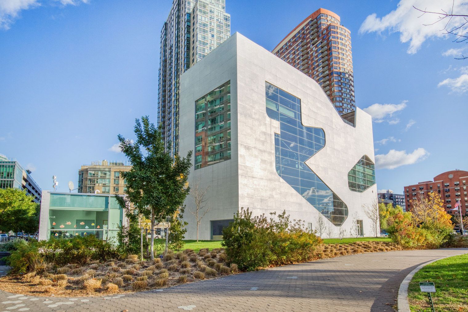 A modern building with a unique angular design, surrounded by other high-rise buildings and a well-maintained park area.