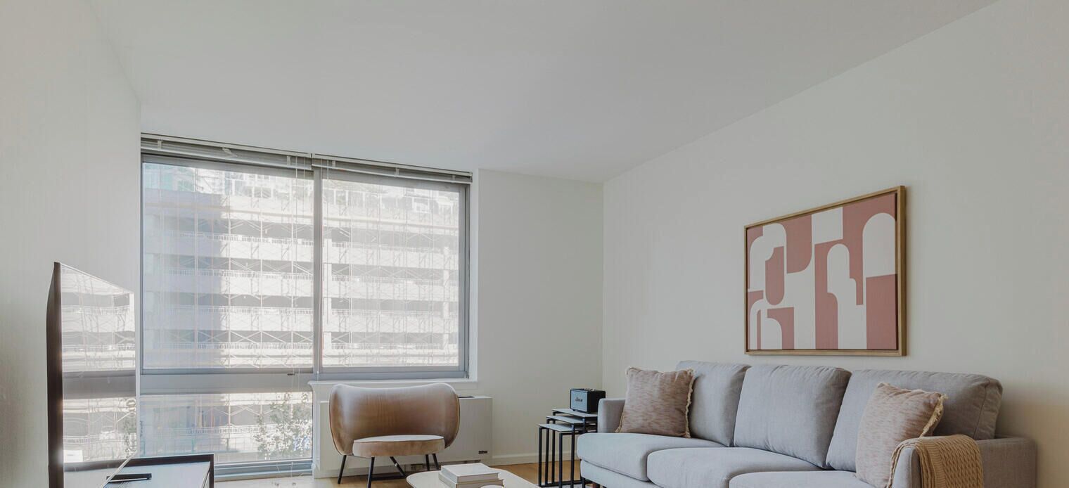 A modern living room with a large window, a sofa, a chair, and a framed abstract artwork.