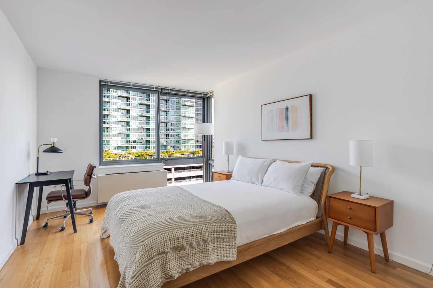 A bedroom with a large bed, a desk, a chair, a bedside table, and a window with a view of buildings.