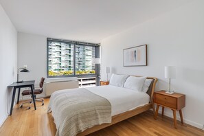 A bedroom with a large bed, a desk, a chair, a bedside table, and a window with a view of buildings. - Blueground | Long Island City, pool, nr Gantry Plaza (New York)
