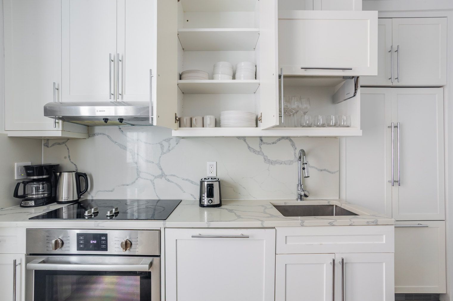 A modern kitchen with white cabinetry, a built-in oven, a coffee maker, a toaster, and a marble backsplash.