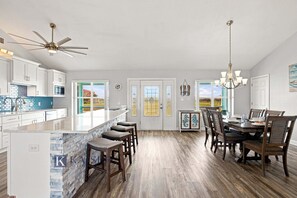 A spacious kitchen with a central island, bar stools, a dining area with a chandelier, and a view through the glass doors. - Little Lagoon Waterfront • Private Pier • West Beach •  Dockside Oasis by Kaiser (Gulf Shores)