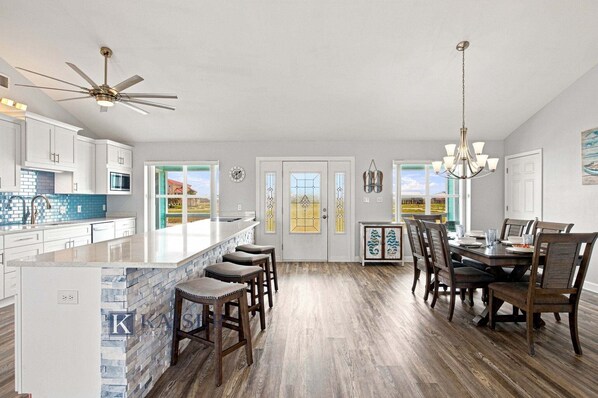 A spacious kitchen with a central island, bar stools, a dining area with a chandelier, and a view through the glass doors. - Little Lagoon Waterfront • Private Pier • West Beach •  Dockside Oasis by Kaiser (Gulf Shores)