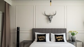 Une chambre d'hôtel avec un lit, deux oreillers ornés de la mention « foyer heureux », un crâne de taureau décoratif, une table de chevet, une lampe de sol et un vase rempli de fleurs.