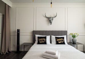 Ein Hotelzimmer mit einem Bett, zwei Kissen mit dem Aufdruck 'happy home', einem dekorativen Stierkopf, einem Nachttisch, einer Stehlampe und einer Vase mit Blumen.