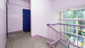 Un couloir avec une porte bleue, une fenêtre donnant sur un paysage verdoyant et une rambarde rose.