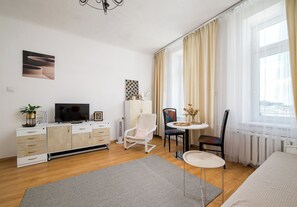 A modern living room with a flat-screen TV, a white cabinet with patterned drawers, a white chair, a small round table, and a large window with sheer curtains. - Hoża Artistic Charm Studio (Warszawa)