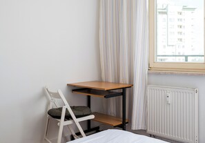 A corner room with a bed, a white chair, a wooden shelf, a radiator, and a window with a view of buildings. - Metro Wilanowska Classic Apartment (Warszawa)