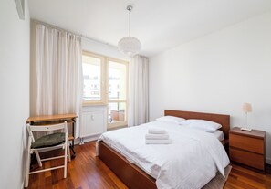 Une chambre à coucher avec un lit en bois, du linge de lit blanc, un bureau avec une chaise, une table de chevet et une lampe.