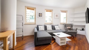 Ein modernes Wohnzimmer mit grauem Sofa, einem Couchtisch und einem Sessel mit einem Kissen mit Mops-Motiv. Zwei Fenster mit Jalousien, eine Wandregal und eine Deckenleuchte sind ebenfalls vorhanden.