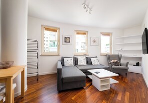 Ein modernes Wohnzimmer mit grauem Sofa, einem Couchtisch und einem Sessel mit einem Kissen mit Mops-Motiv. Zwei Fenster mit Jalousien, eine Wandregal und eine Deckenleuchte sind ebenfalls vorhanden.