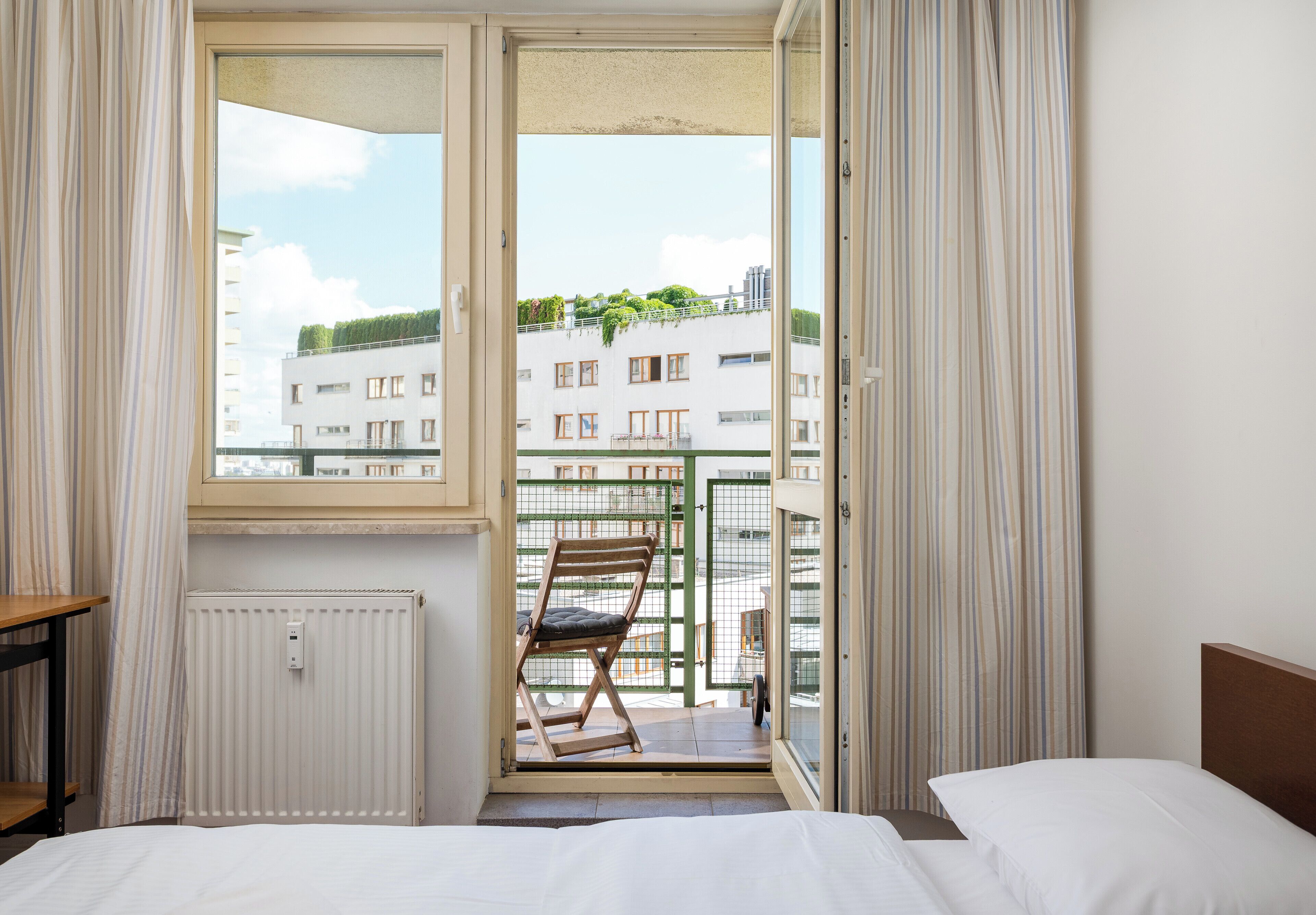 A hotel room with a bed, a chair, a radiator, and a view of buildings through an open door.