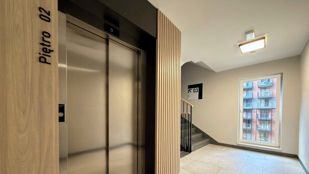 An interior corridor with a wooden door labeled 'Pietro 02,' a staircase, and a window with a view of a building. - Madalińskiego Modern Studio 8B (Wrocław)