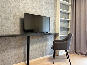 Une chambre moderne avec un téléviseur à écran plat posé sur un meuble noir, une chaise grise et une étagère blanche adossée à un mur gris texturé.