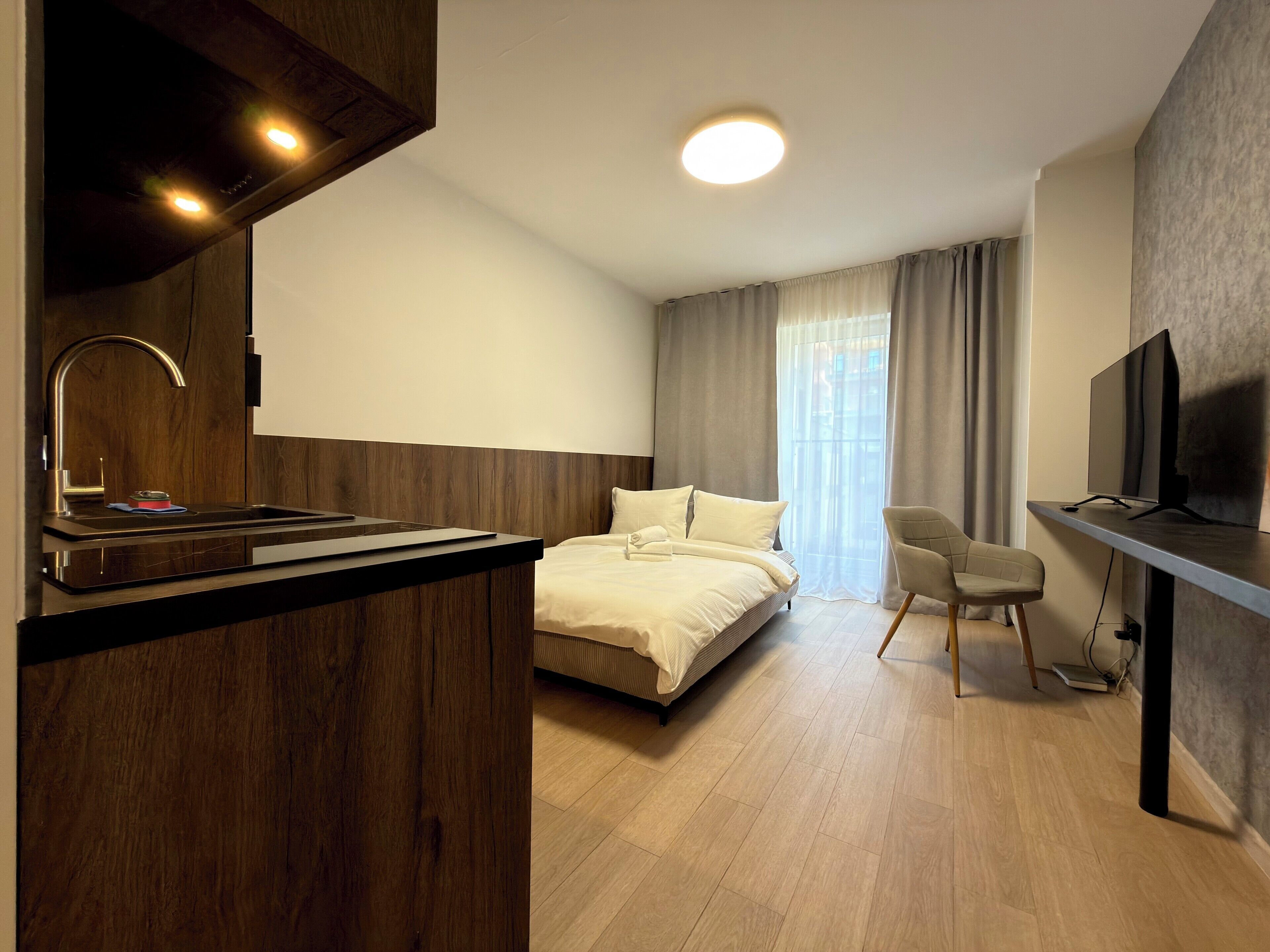 Una habitación de hotel moderna con una kitchenette de madera, una cama con ropa blanca, un televisor y un ventanal con cortinas.