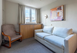 A room with a wooden chair, a light-colored sofa, a window with curtains, and a wall-mounted lamp.