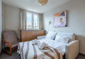 A hotel room with a bed, a chair, a desk, a window with curtains, and a painting on the wall. - Szczecin City Center Apartment (Szczecin)