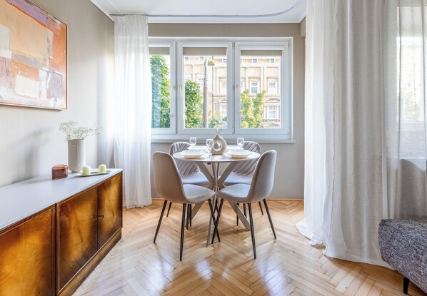 A dining area with a table set for four, a wooden herringboard, and a view of buildings through a window with white curtains.