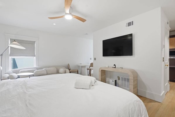 A modern hotel room with a large bed, a flat-screen TV mounted on the wall, a ceiling fan, a small round table, and a seating area with cushions. - The Palmetto Suite at One 01 (Savannah)