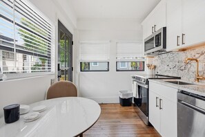 Een kleine keuken met witte kasten, een magnetron, oven en een spoelbak. Er staat een eettafel met een stoel, en een raam met jaloezieën.