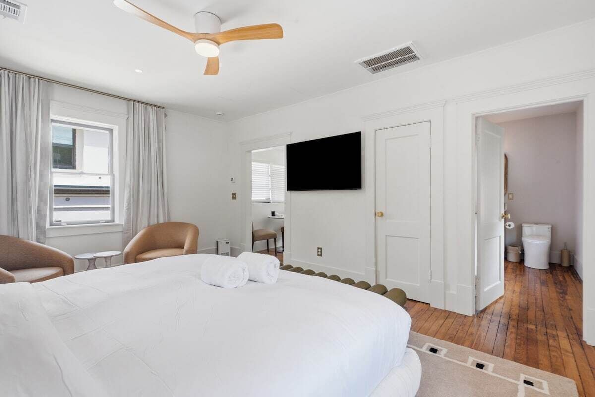 A hotel room with a large bed, a ceiling fan, a flat-screen TV, a window with curtains, a door leading to a bathroom, and a small table with chairs.