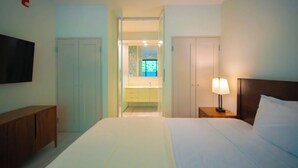 Ein Hotelzimmer mit Bett, Nachttischlampe, an der Wand montiertem Fernseher, Holzschrank und einer Tür zu einem Badezimmer mit Spiegel und Waschbecken.