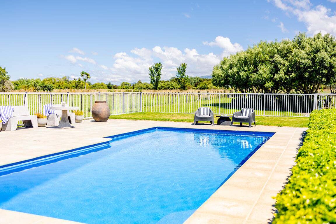 A rectangular swimming pool with a tiled edge, surrounded by a white fence, and a grassy area with trees and shrubs.