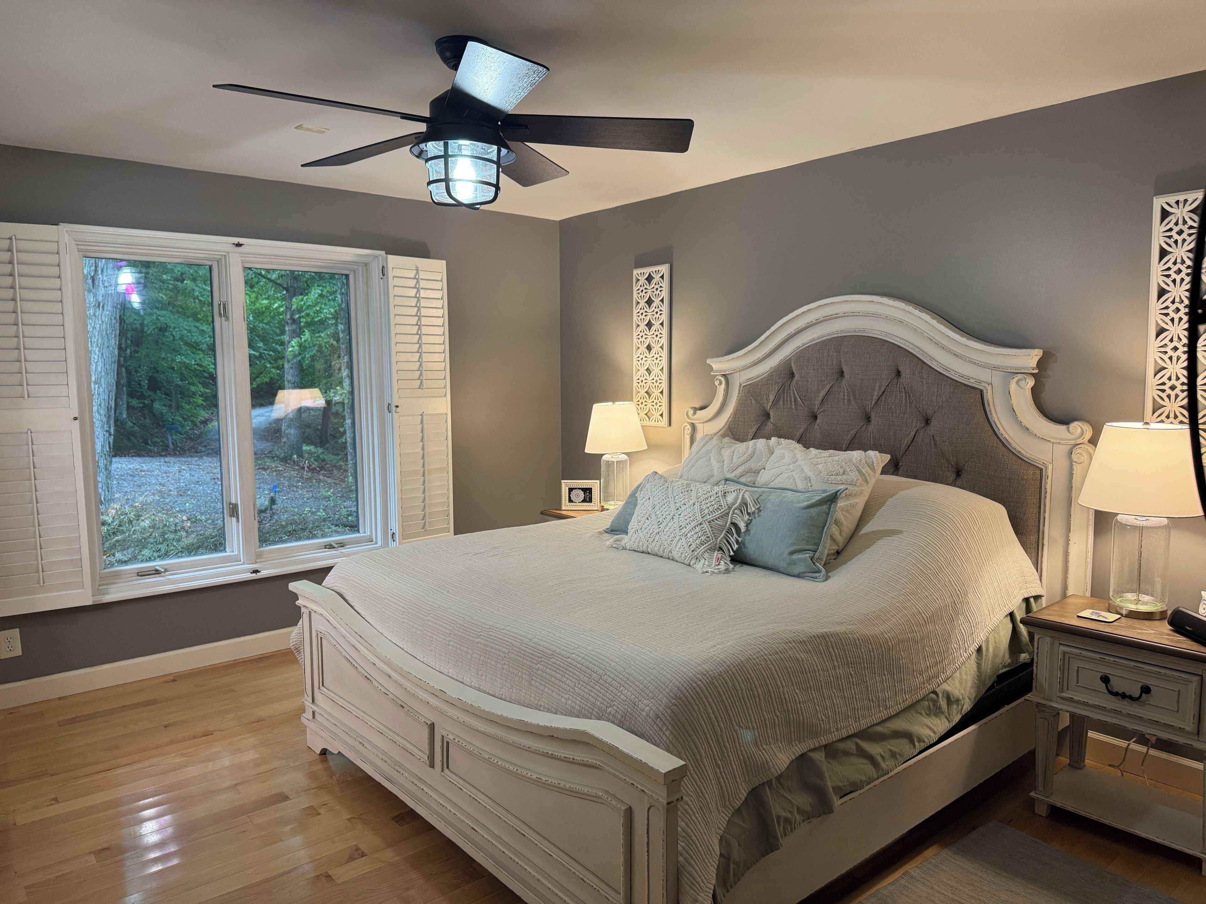 Une chambre à coucher avec un grand lit, un ventilateur de plafond, une fenêtre avec des volets et une table de chevet avec une lampe.