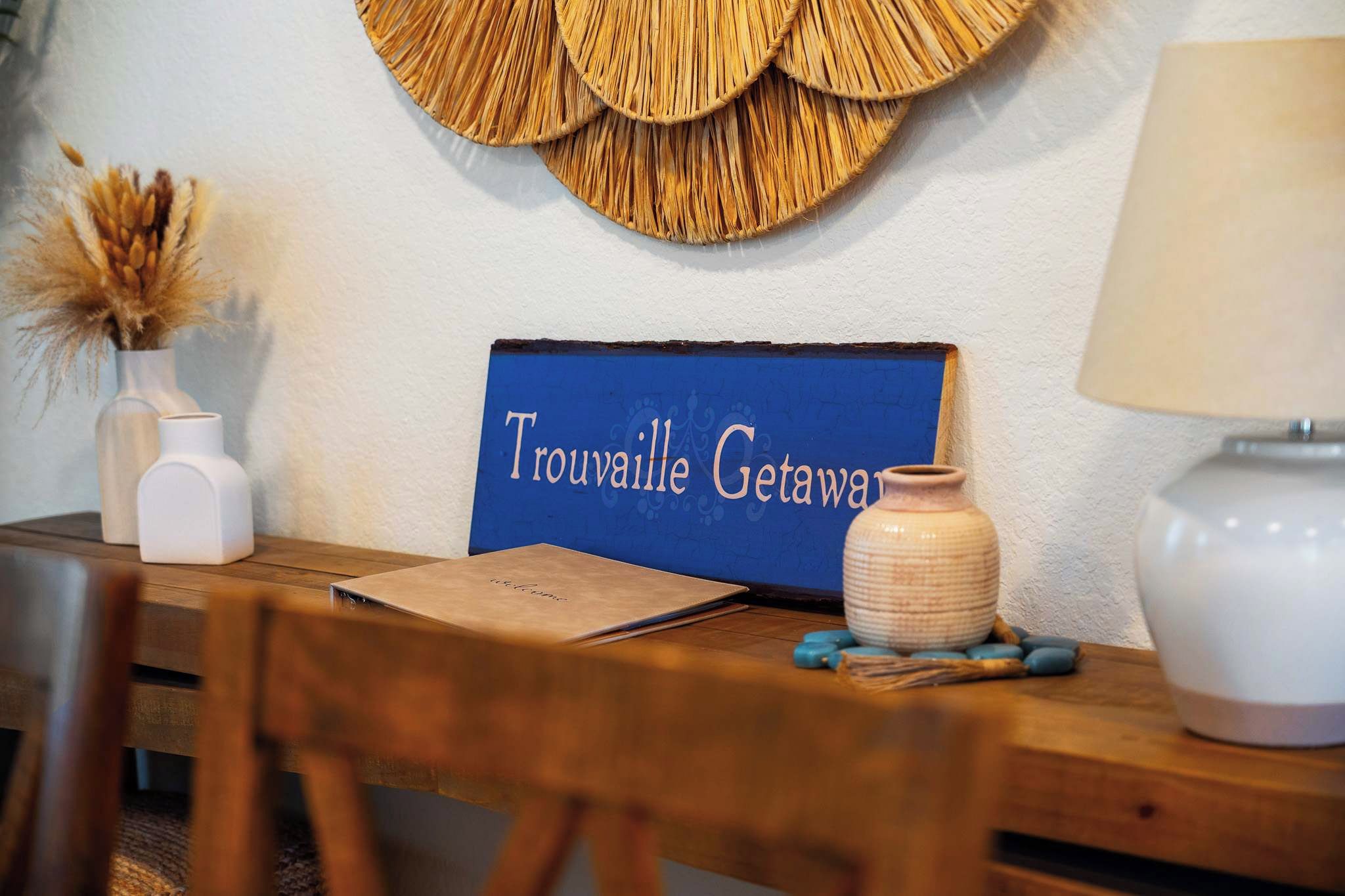 A wooden table with a sign reading 'Trouville Getaway,' a vase with dried plants, a lamp, and a notebook.