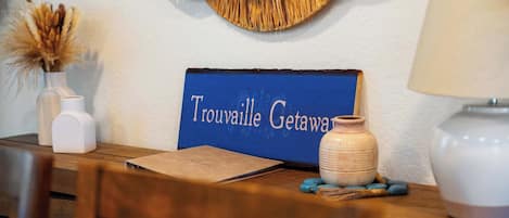 A wooden table with a sign reading 'Trouville Getaway,' a vase with dried plants, a lamp, and a notebook.
